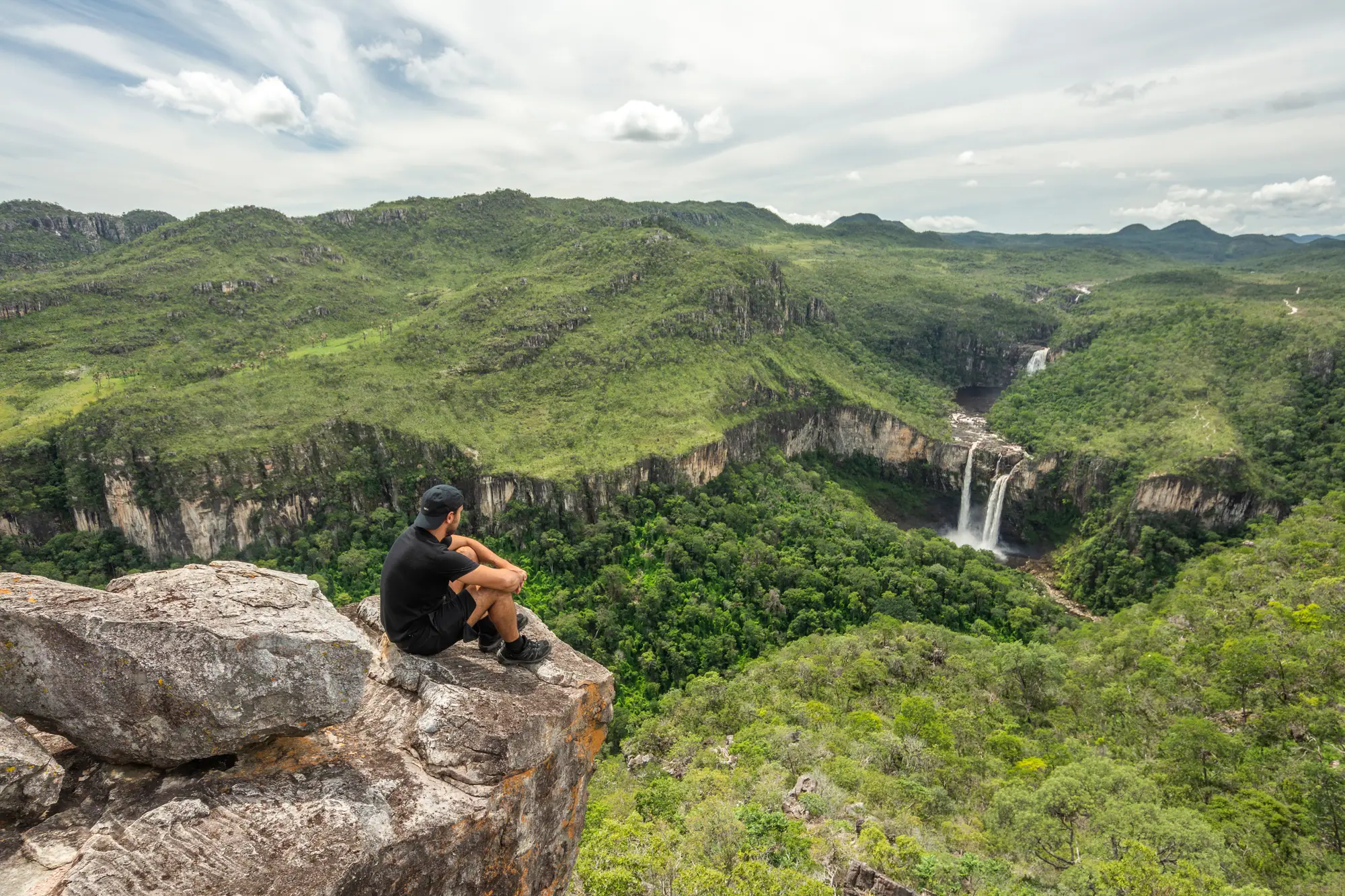 Hiker on a rocky ledge overlooks a green Cerrado canyon with twin waterfalls under soft, cloudy skies.