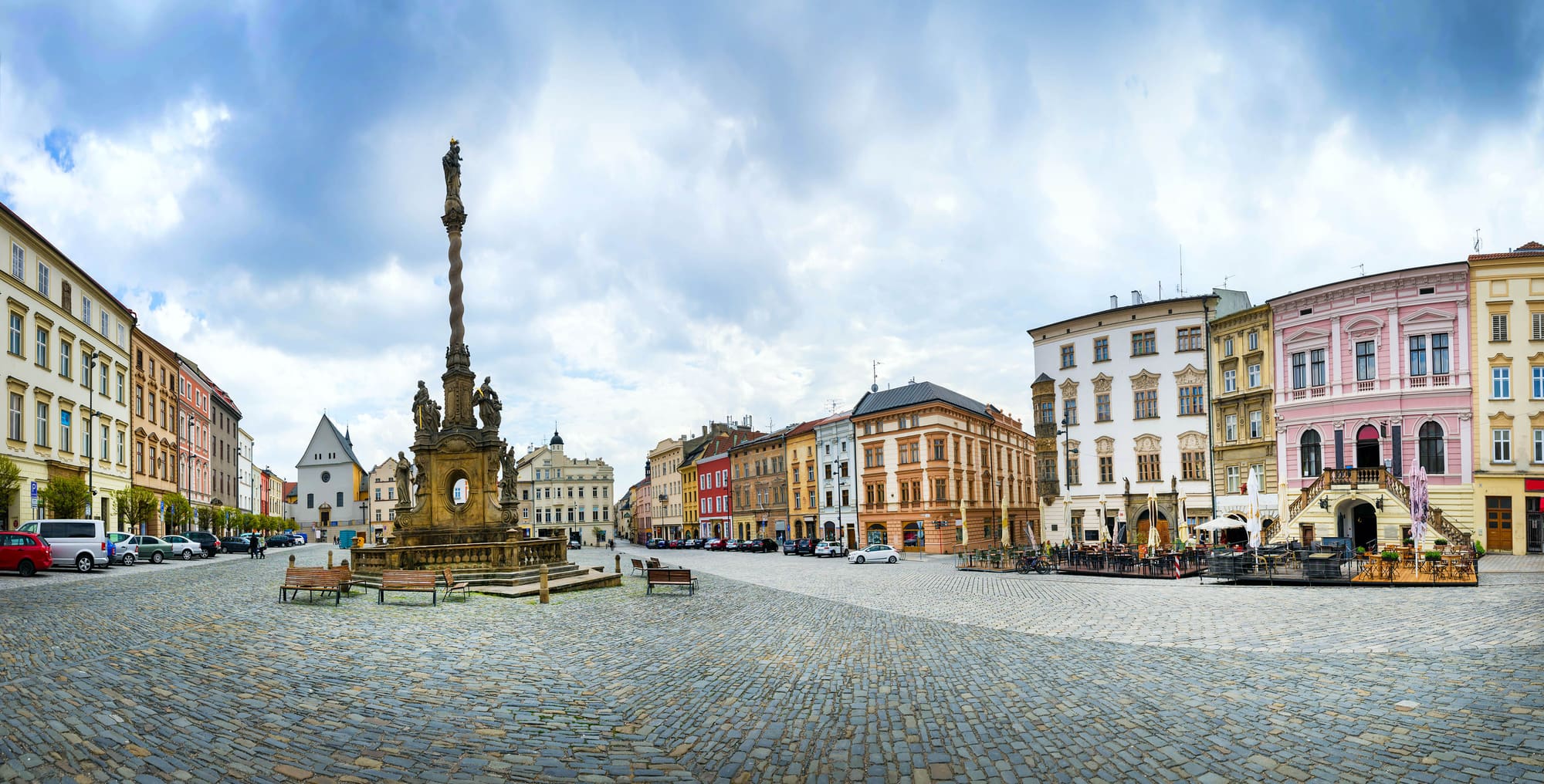 Baroque Holy Trinity Column rises above a cobbled square in Olomouc, flanked by pastel townhouses under cloudy skies.
