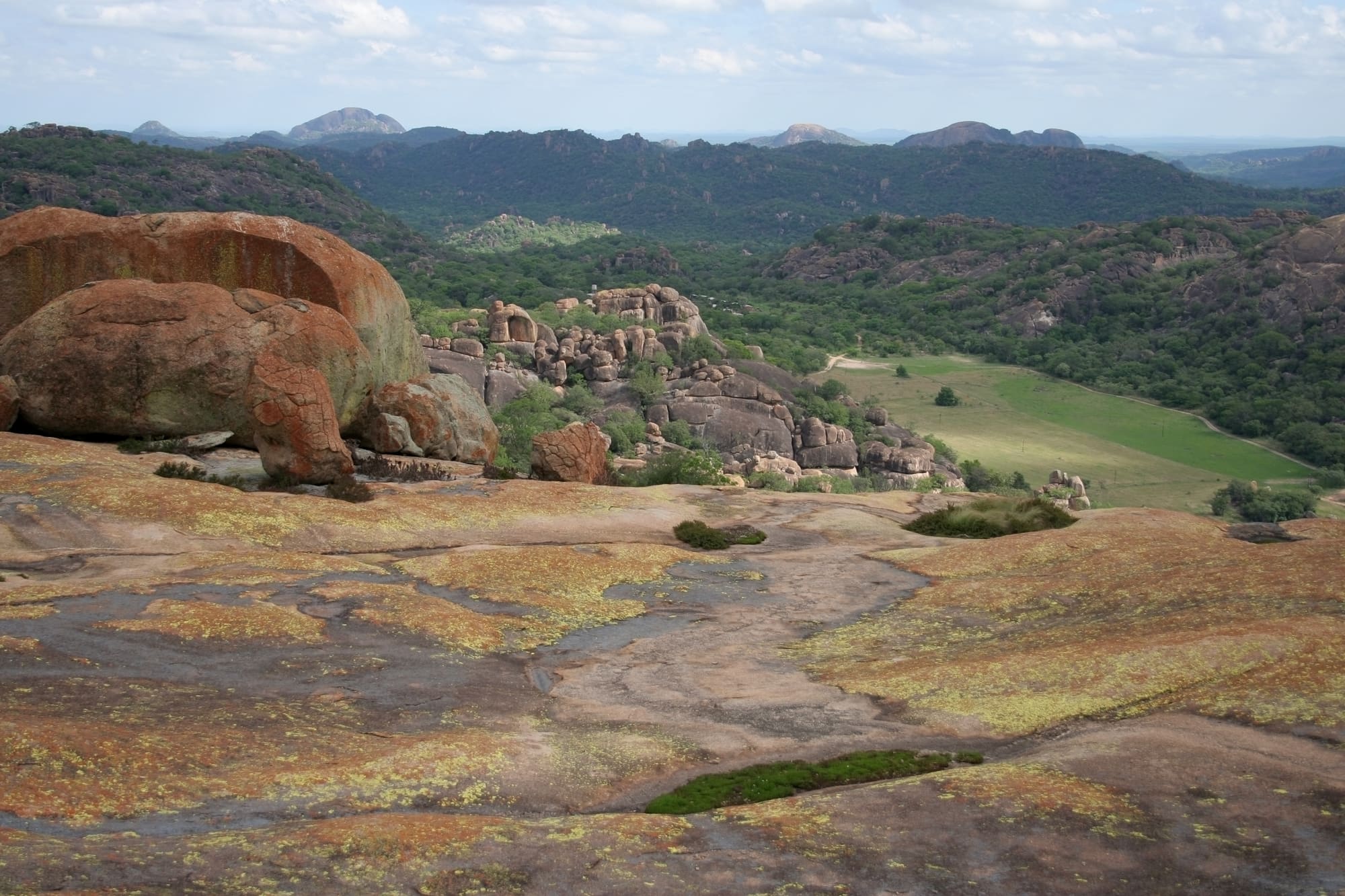 Rounded granite boulders stained orange with lichen overlook a wooded valley in the Matobo Hills under pale sky.
