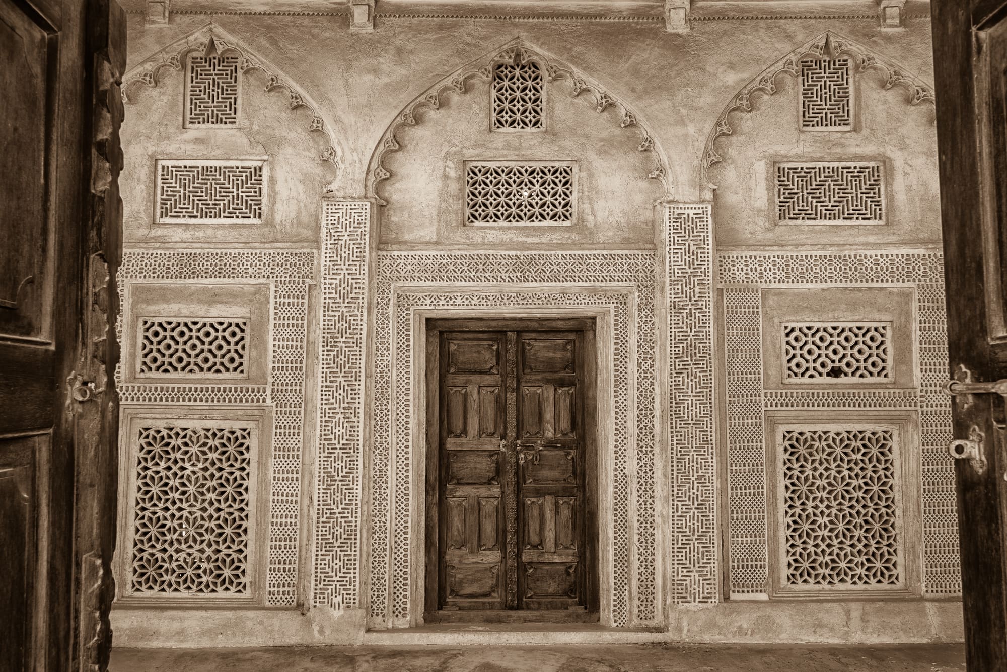 Sepia-toned view of an arched plaster wall with intricate latticework windows and a carved wooden double door.