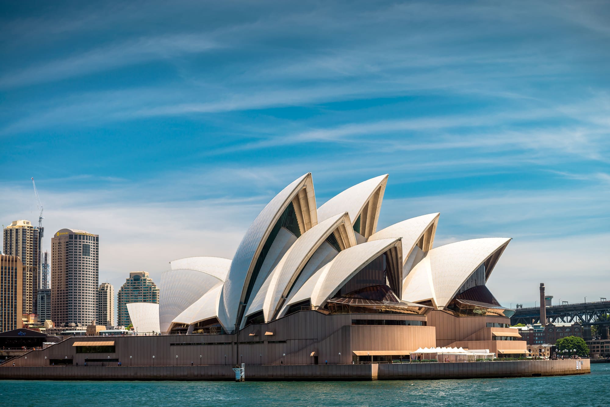 Sydney Opera House's white, sail-like shells rise above harbour water beneath a bright blue sky with wispy clouds.