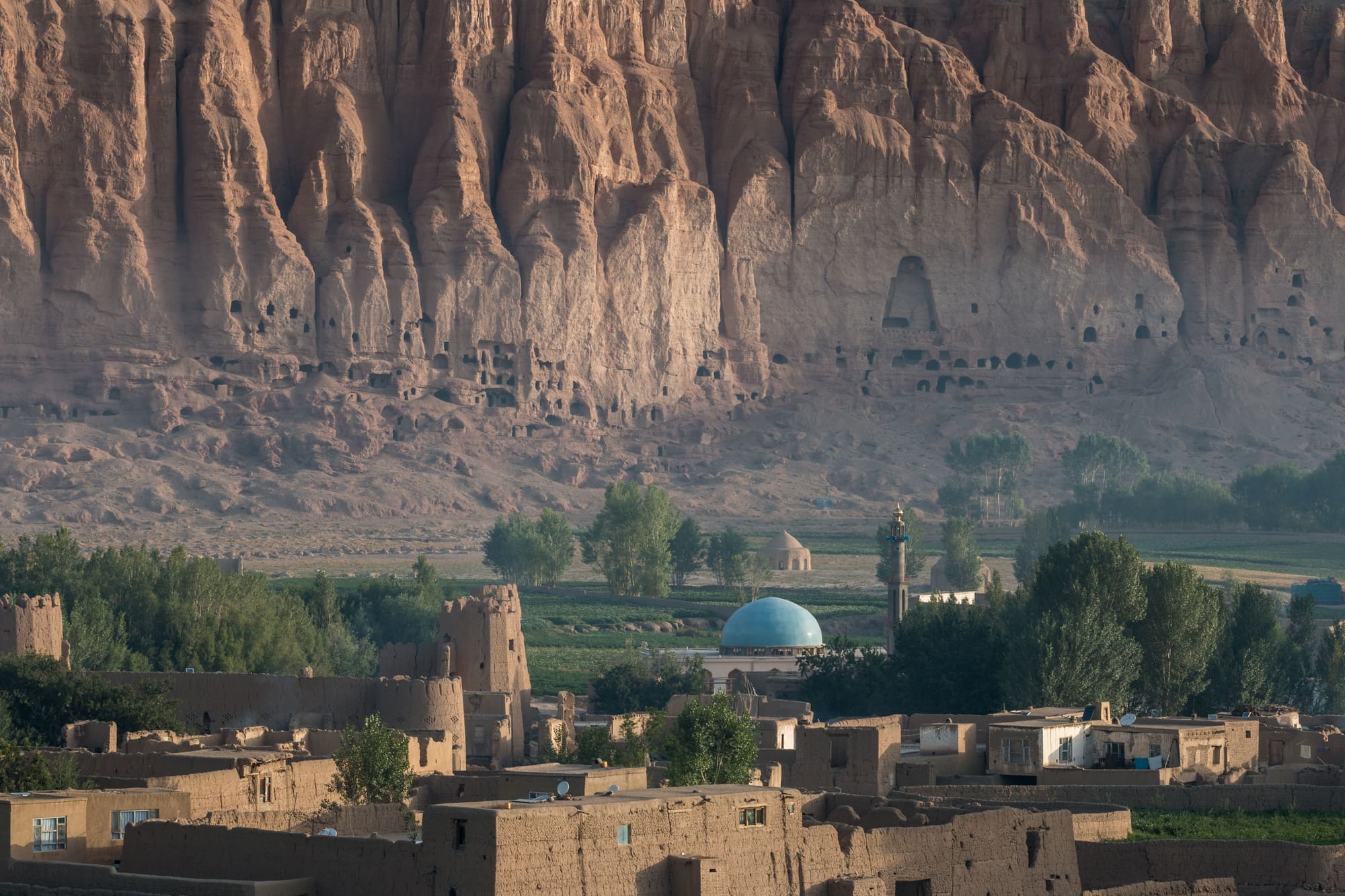 Sunlit cliffs of Bamiyan Valley, honeycombed with caves and niches, rise above a mud-brick village and blue-domed mosque.