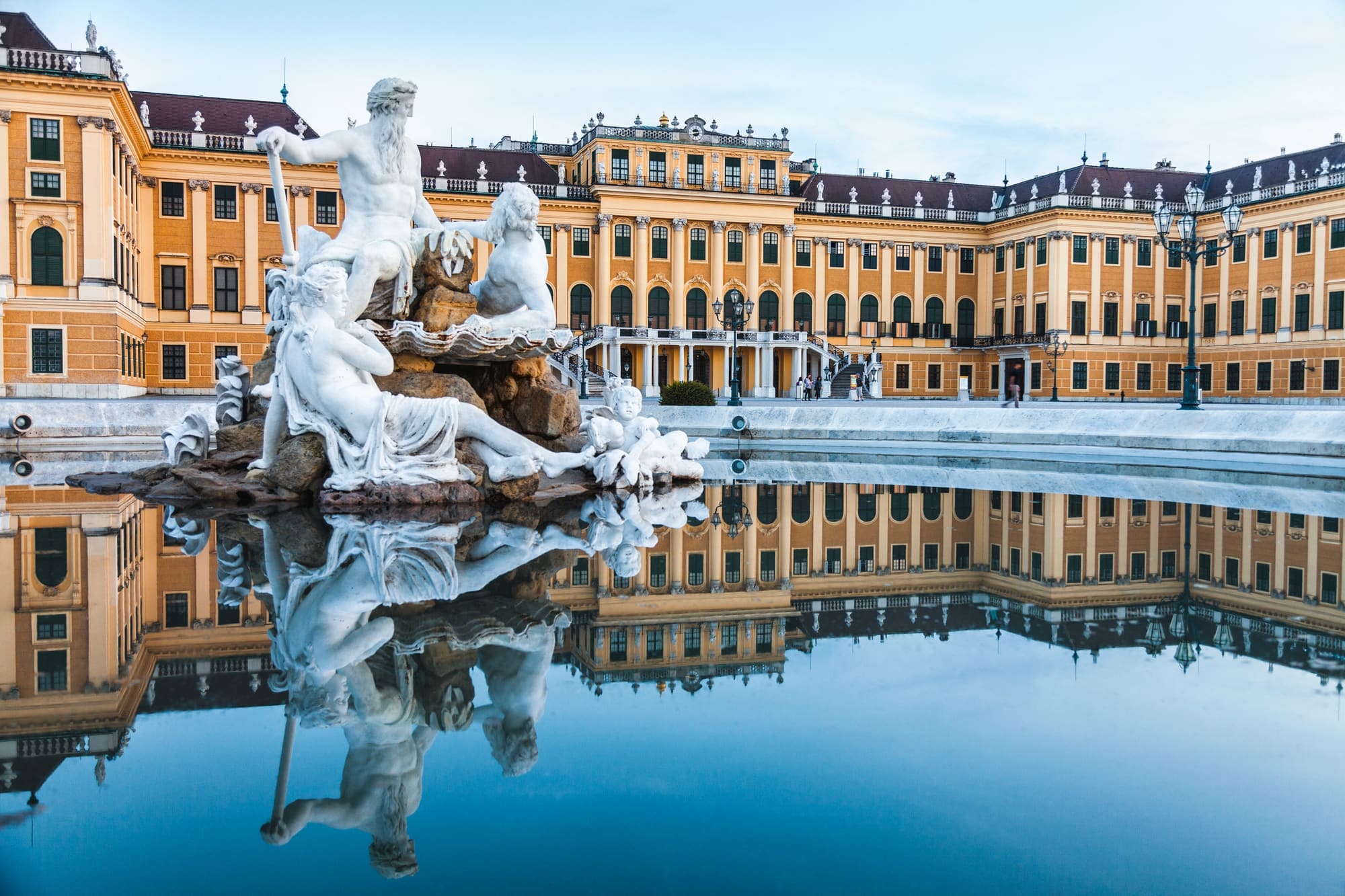 The yellow facade of Schönbrunn Palace, with a fountain of white statues reflected in the pool